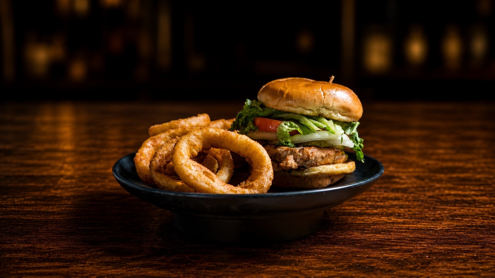 Crispy chicken sandwich with onion rings on a black plate at the bar
